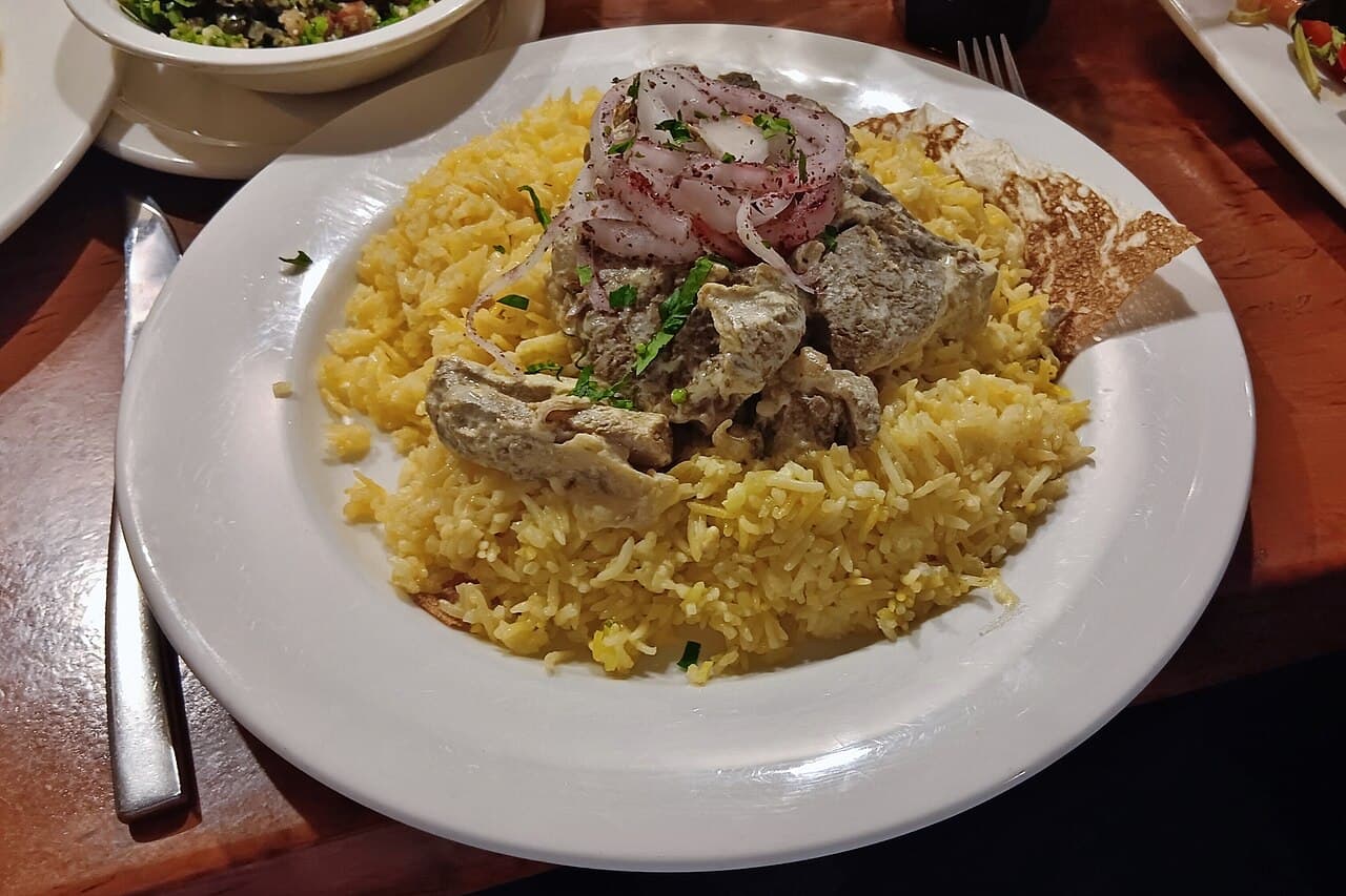A large shared platter of mansaf: lamb and rice topped with almonds, with yoghurt sauce alongside