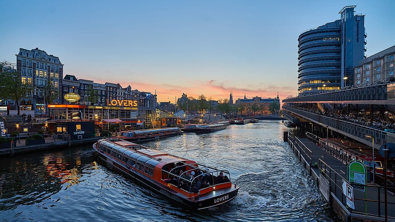 A small boat passing lit canal houses and a bridge at dusk